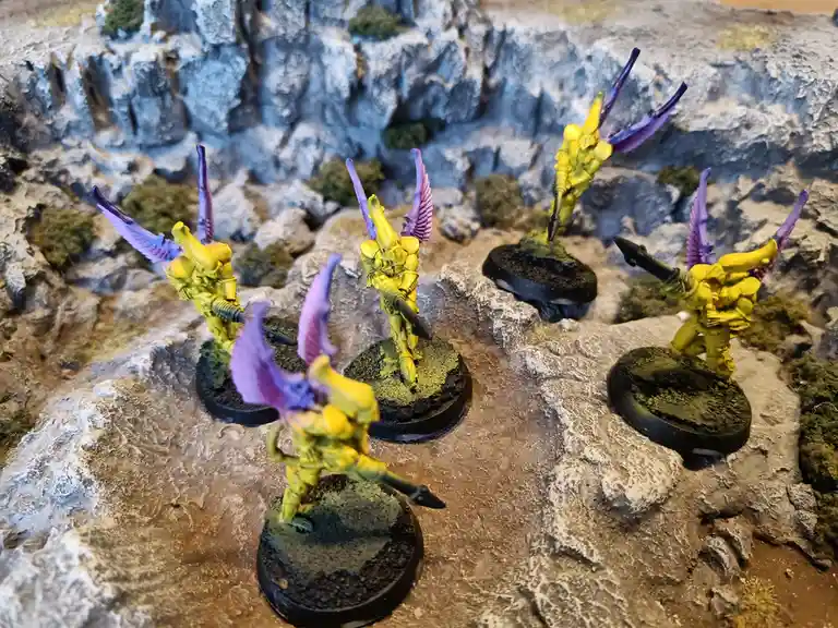 Five yellow-painted miniature figures with purple wings stand on rocky terrain, each holding a weapon. The overlaid text…