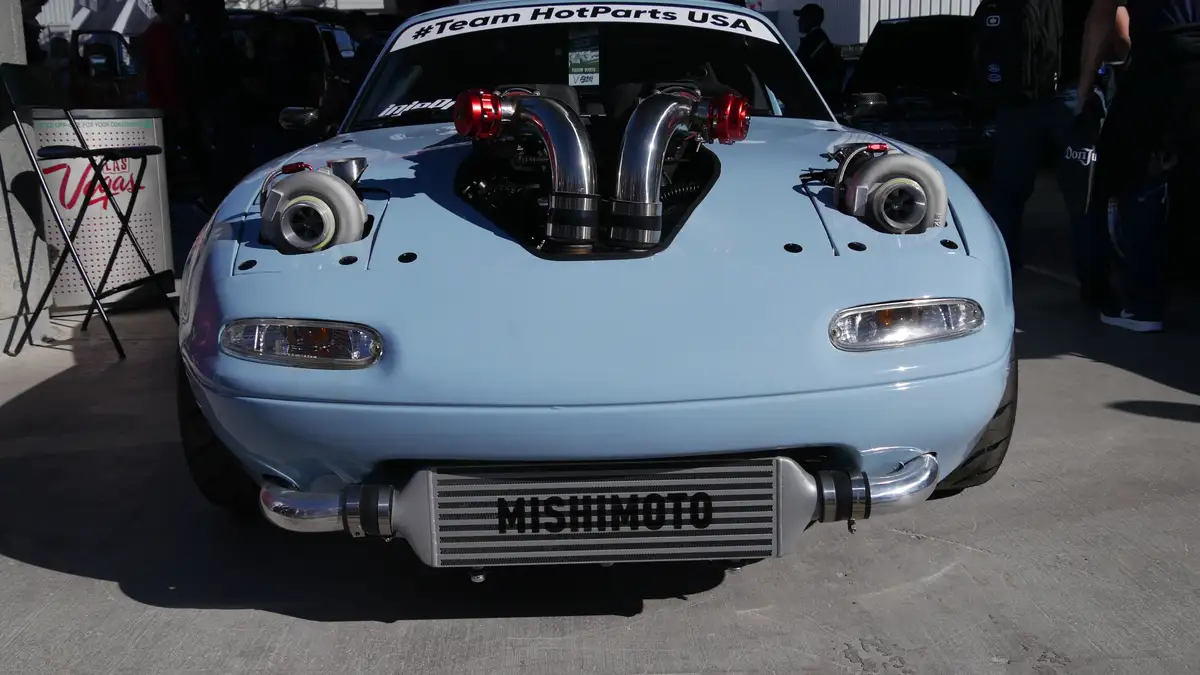 A light blue modified Mazda Miata with its hood removed, revealing a highly customized engine setup with two large…