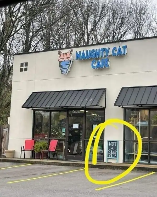 An exterior shot of a building labeled 'NAUGHTY CAT CAFE' with a cartoon cat wearing a face mask above the entrance. A large…