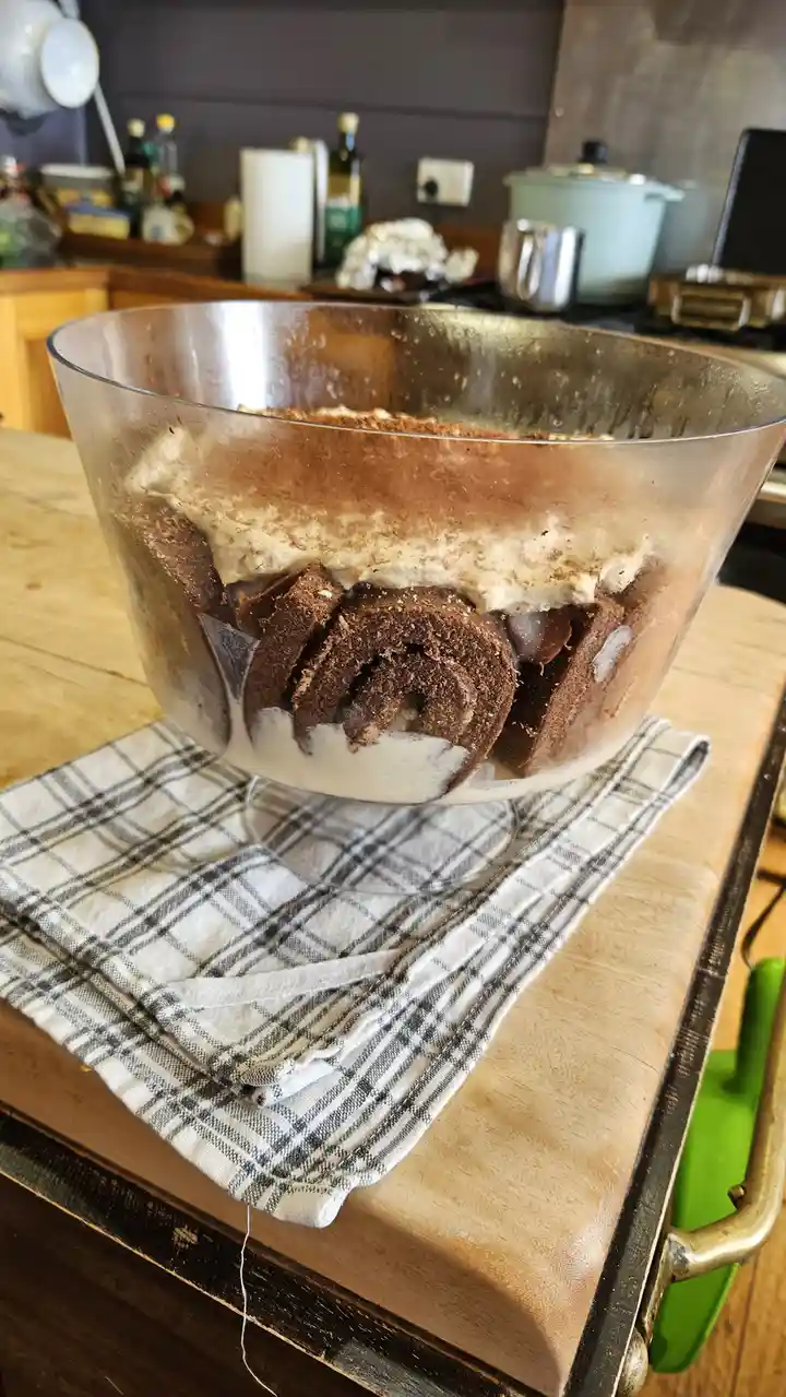 A large glass trifle bowl sits on a wooden counter over a plaid cloth, filled with layers of chocolate cake, cream, and…