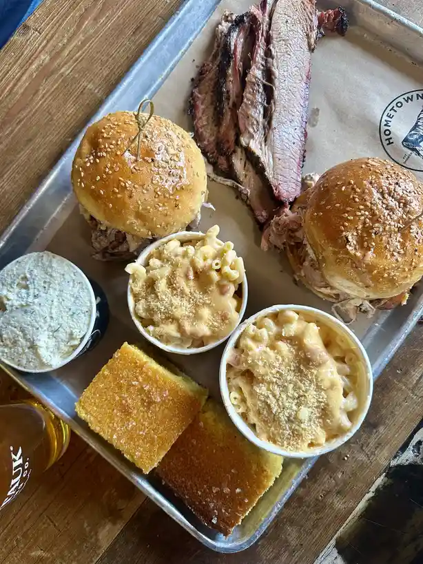 A metal tray holds a hearty meal featuring two pulled pork sliders with sesame seed buns, a large portion of sliced smoked…