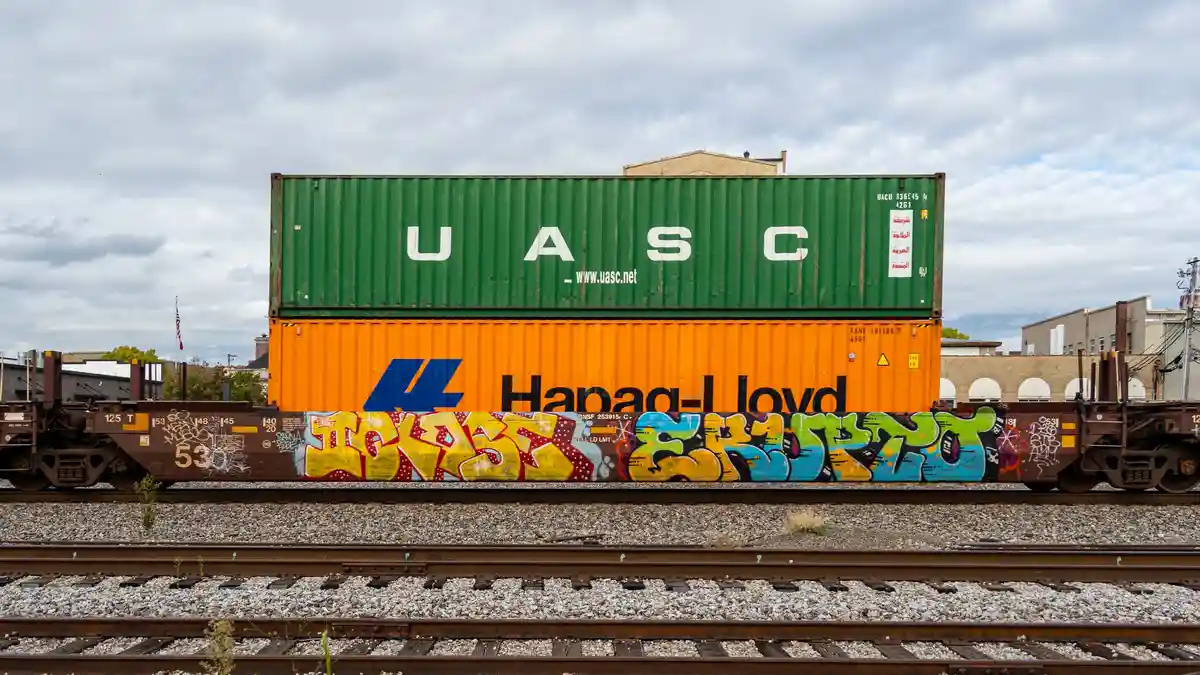 A photograph of two stacked shipping containers on a freight train, the top green one labeled 'UASC' and the bottom orange…