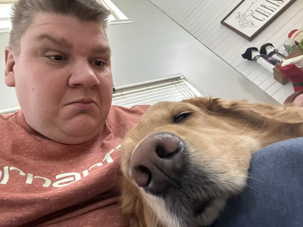 A man with a disgruntled, pouting expression sits next to a sleeping golden retriever whose nose is close to the camera. The…