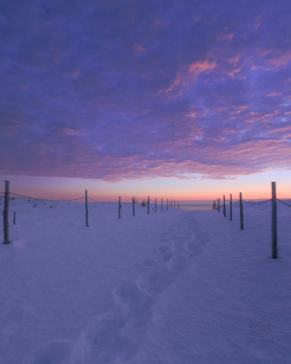 A serene snowy path with footprints leading toward a horizon under a vibrant purple and pink sunset sky, flanked by a rope…