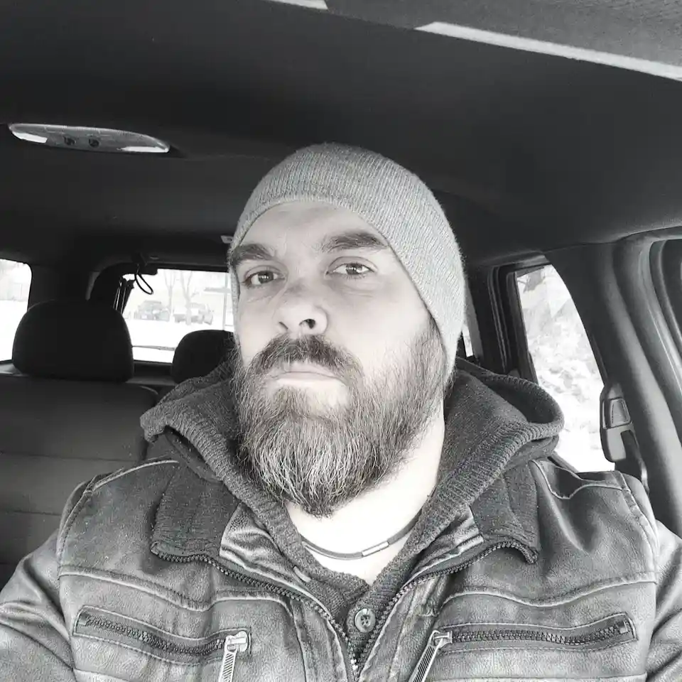 A black-and-white selfie of a bearded man wearing a beanie and a leather jacket, sitting inside a car with snow visible…