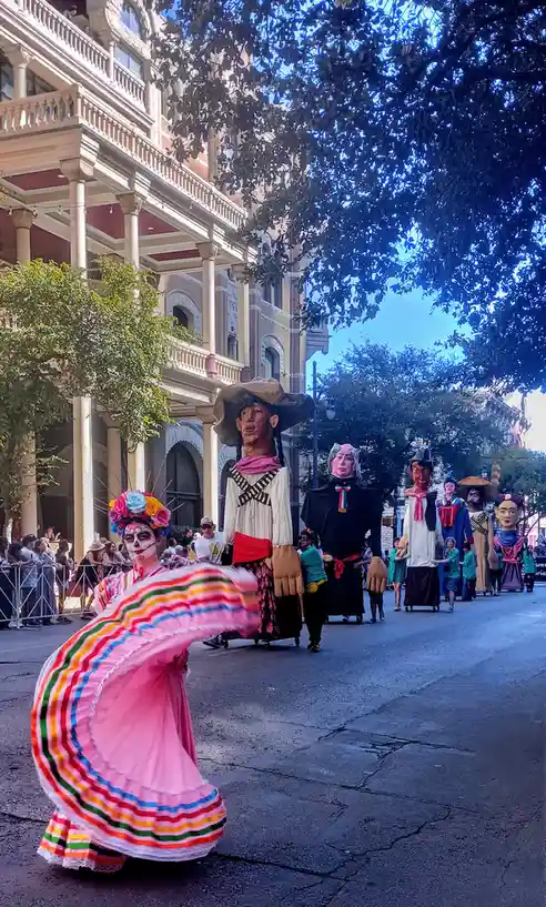 A street parade features a dancer in a colorful, flowing pink dress with sugar skull makeup and a floral headdress, spinning…