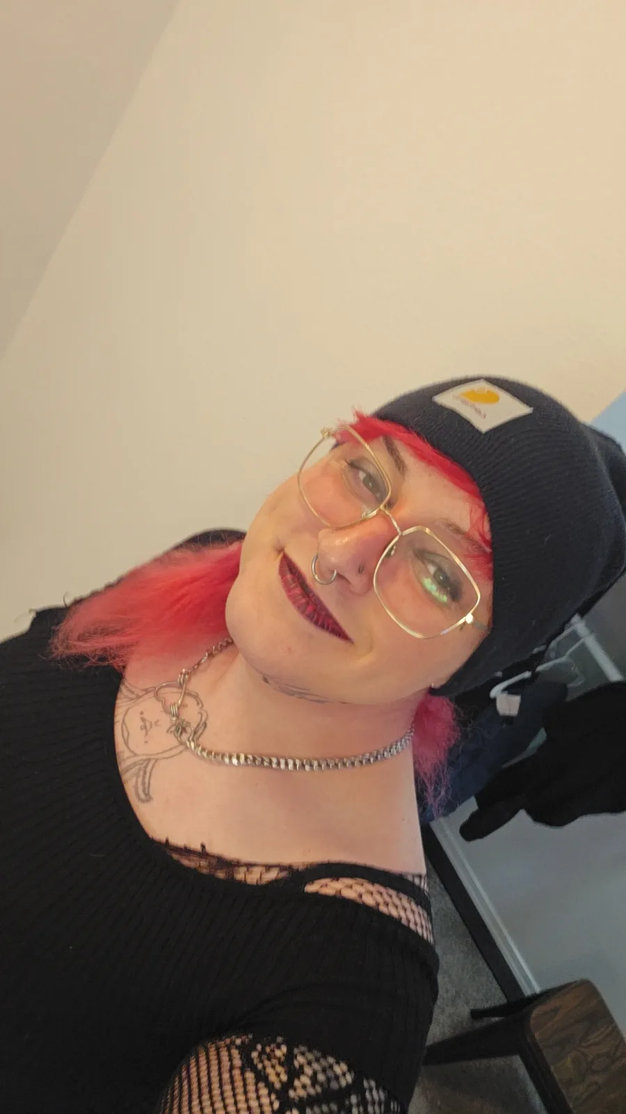 A person with bright pink hair, wearing a black Carhartt beanie, gold-rimmed glasses, and a black top with lace details…