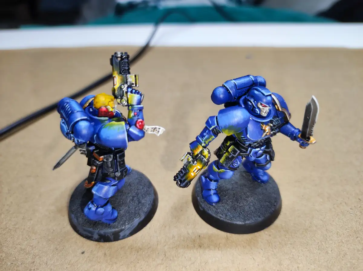 Two blue-painted miniature space marines in power armor stand on black circular bases on a brown surface. The figure on the…