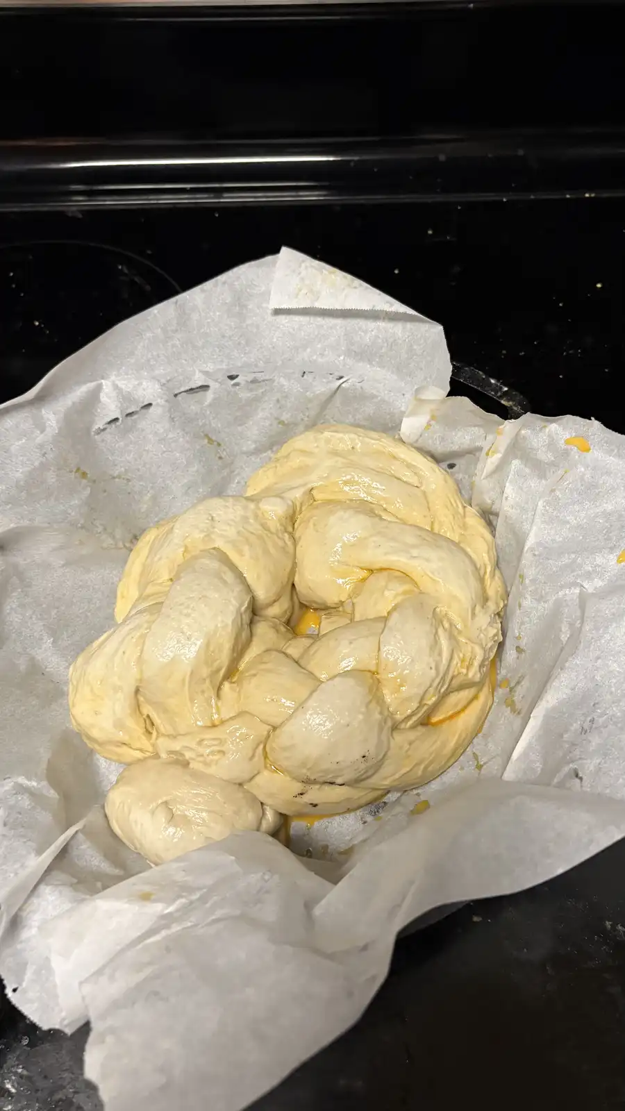 A close-up photo shows a large, pale yellow, braided dough loaf resting on parchment paper inside a dark baking pan. The…