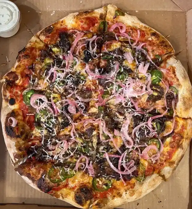 An overhead photo of a round pizza in a cardboard box, topped with charred mushrooms, green jalapeños, and thinly sliced…