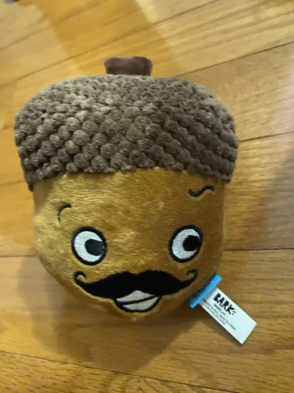 A plush toy shaped like an acorn with a brown, textured cap and a tan body, featuring large cartoon eyes, a smiling mouth…