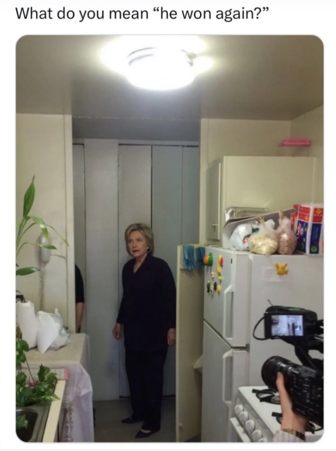 A woman with short blonde hair, wearing a dark suit, stands in a kitchen doorway looking surprised or confused. A white…