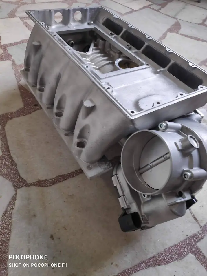 A close-up photo of a silver car engine block and throttle body assembly resting on a tiled floor. The overlaid text reads…