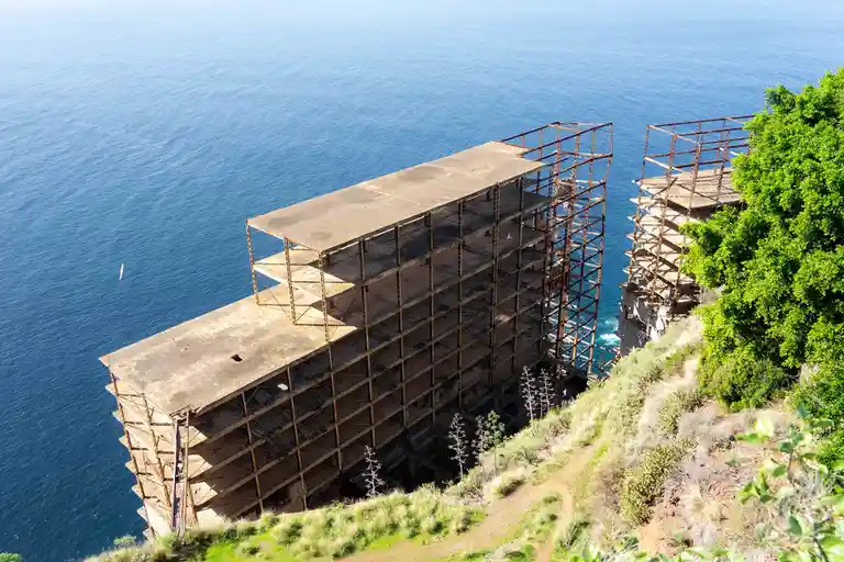 an aerial view shows a partially constructed, skeletal concrete building perched on a cliffside overlooking a vast blue…