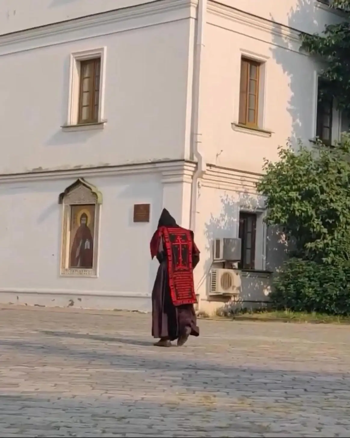 A person in dark robes with a large red patterned vestment walks away from the camera on a paved courtyard. On the white…