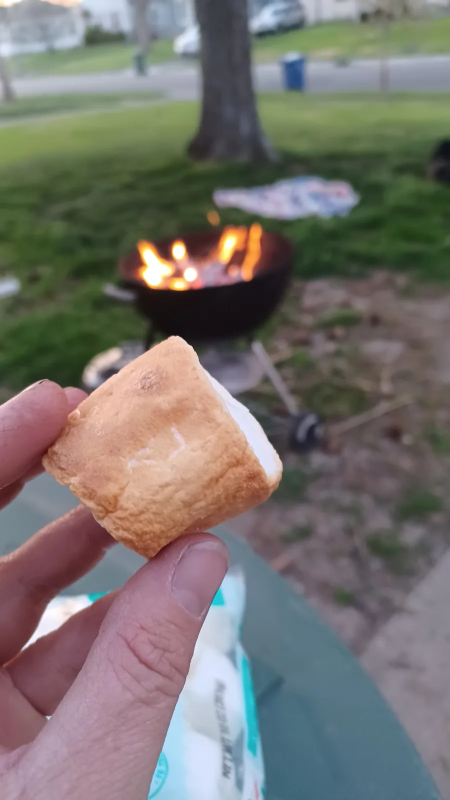 A hand holds up a perfectly toasted square marshmallow with golden-brown edges, with a fire pit burning in the blurred…