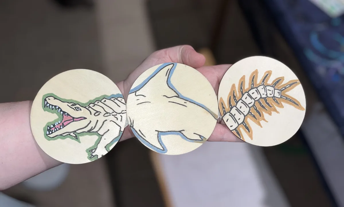 A hand holds three round wooden discs, each painted with a simple outline drawing of a different creature: an alligator on…