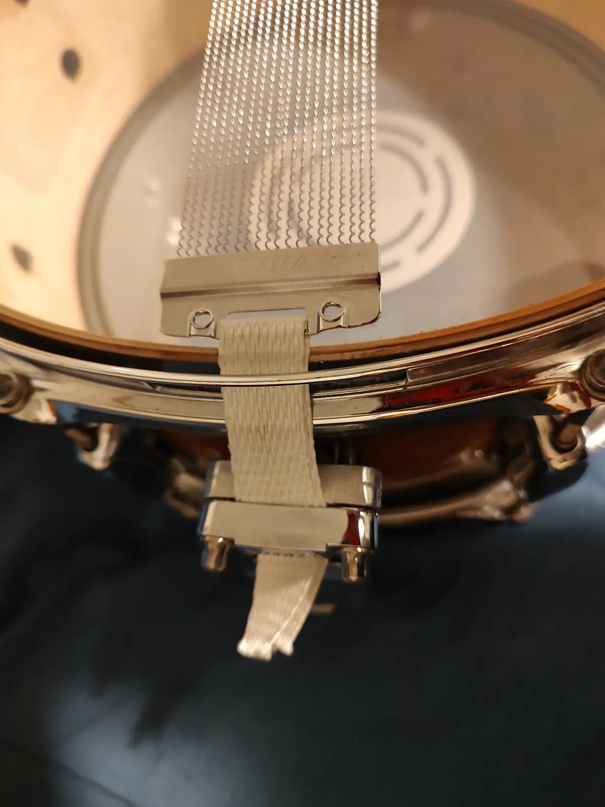A close-up photo of a snare drum's hardware, showing a white fabric strap attached to a chrome bracket holding a mesh snare…
