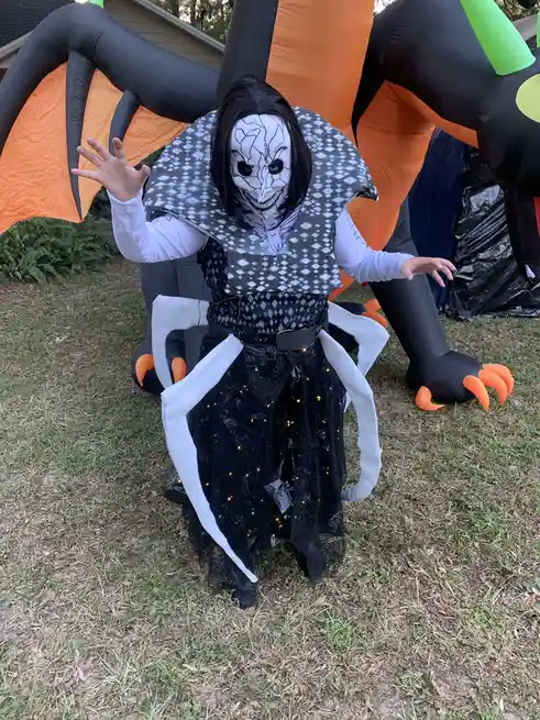 A person in a Halloween costume featuring a white cracked-face mask, black and white patterned cape, and a black skirt with…