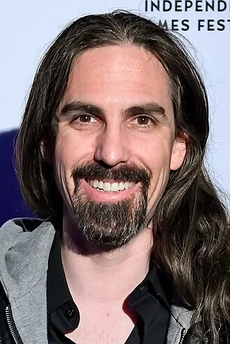 A man with long dark hair, a goatee, and a wide smile is photographed at an event. Behind him, partially visible text reads…