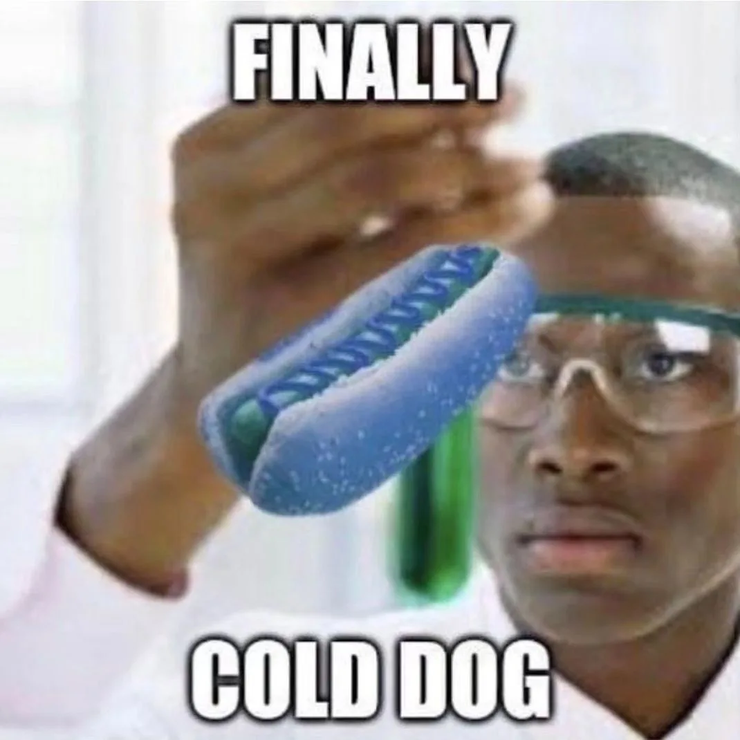 A man wearing safety goggles and a white lab coat holds up a blue, frost-covered hot dog in a test tube-like holder, looking…
