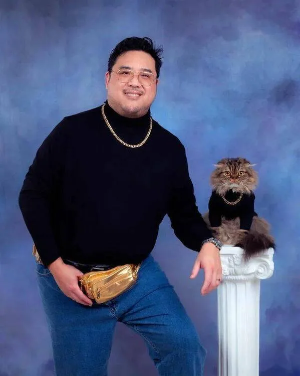 A man in a black turtleneck, gold chain, and gold belt bag poses with a fluffy cat wearing a matching gold chain, both…