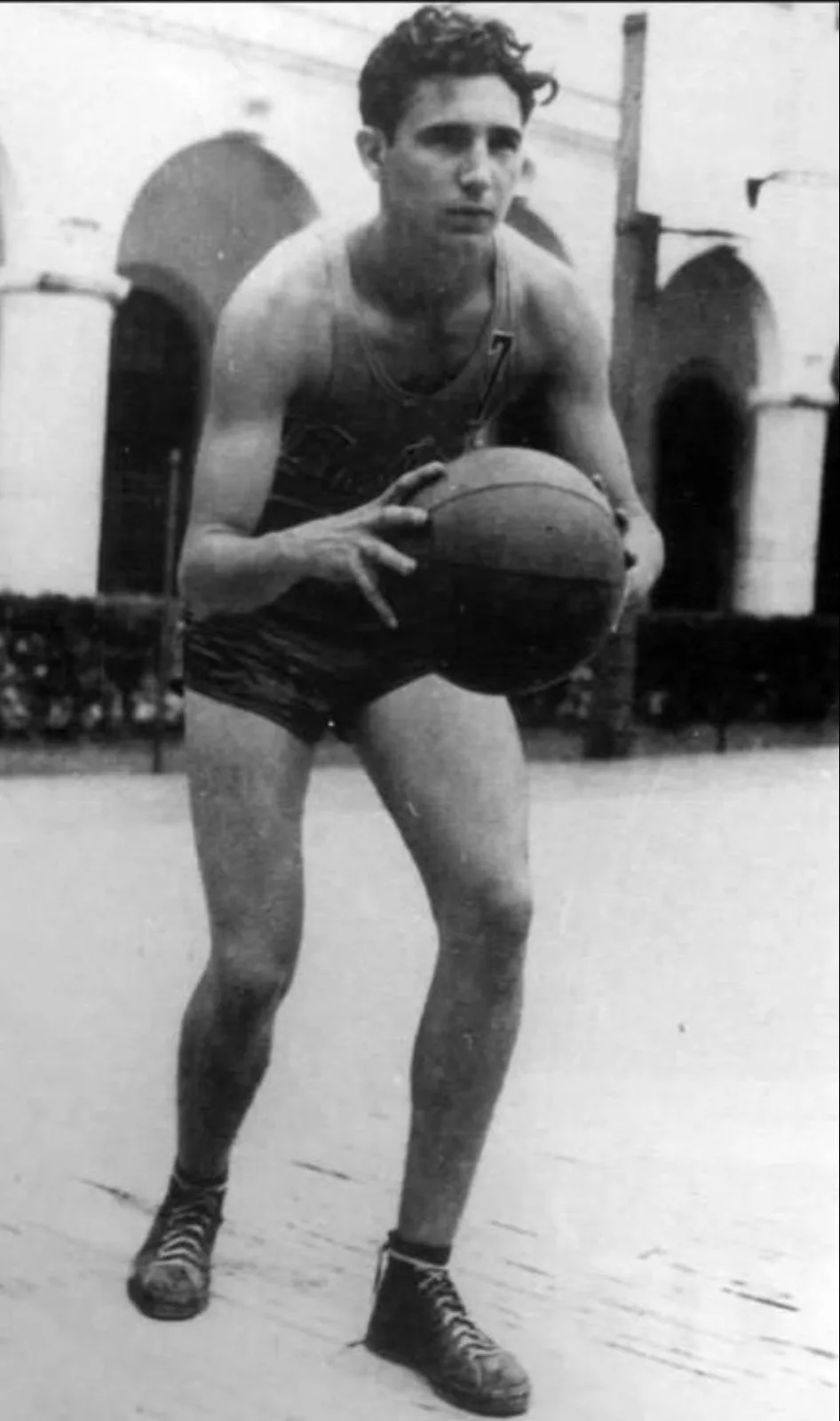 A black-and-white photo of a young man in vintage basketball gear holding a ball in a low stance, with overlaid text reading…