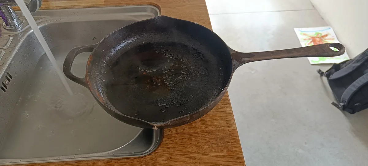 A well-used, dark cast iron skillet sits partially in a stainless steel kitchen sink with running water, its surface…