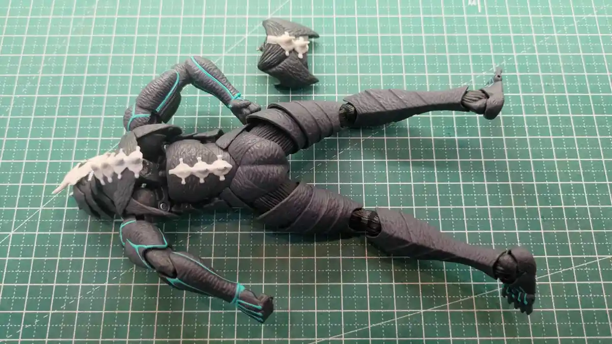 A black action figure with teal accents lies sprawled on a green cutting mat, its head detached and placed nearby. White…