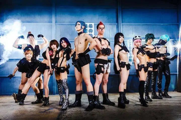 A group of nine people in stylized, revealing black outfits pose confidently in front of a blue corrugated metal wall with…