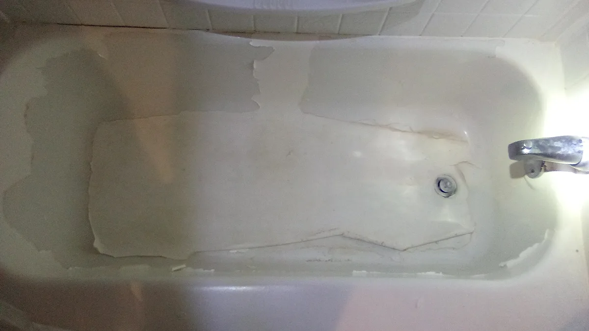 A top-down view of a white bathtub with visible wear, peeling paint, and a chrome faucet on the right. The overlaid text…