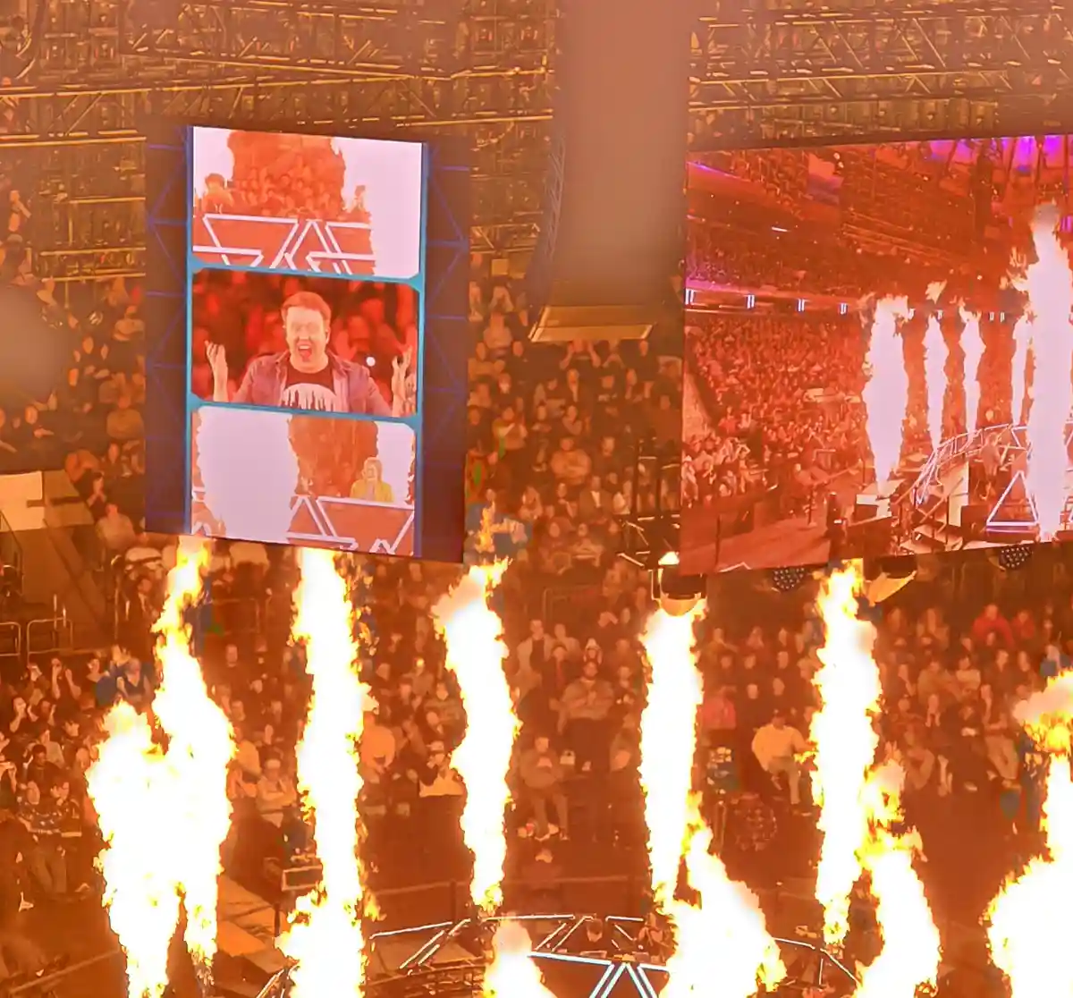 A concert venue with multiple large screens showing a crowd and a man with an excited expression, surrounded by multiple…