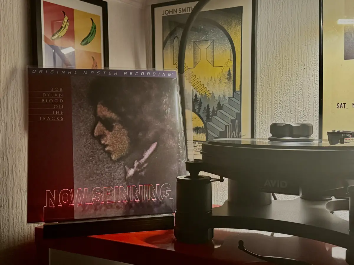 A vinyl record sleeve for Bob Dylan's 'Blood on the Tracks' is propped up next to a turntable, with the text 'NOW SPINNING'…