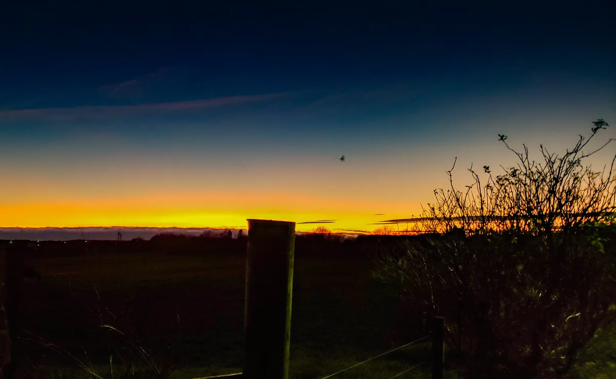 A serene twilight landscape with a vibrant orange and yellow horizon fading into a deep blue sky, framed by dark silhouettes…