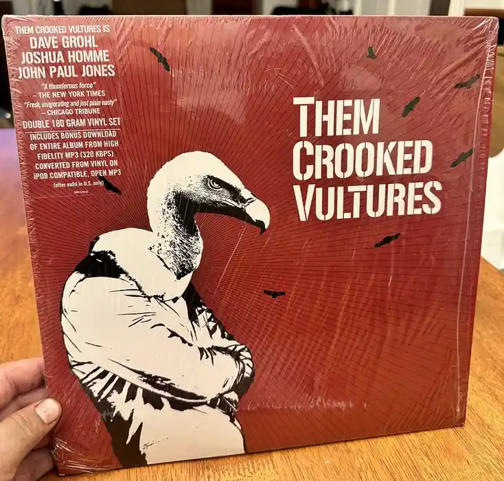 A hand holds a sealed red vinyl record sleeve for 'Them Crooked Vultures' featuring a black-and-white illustration of a…