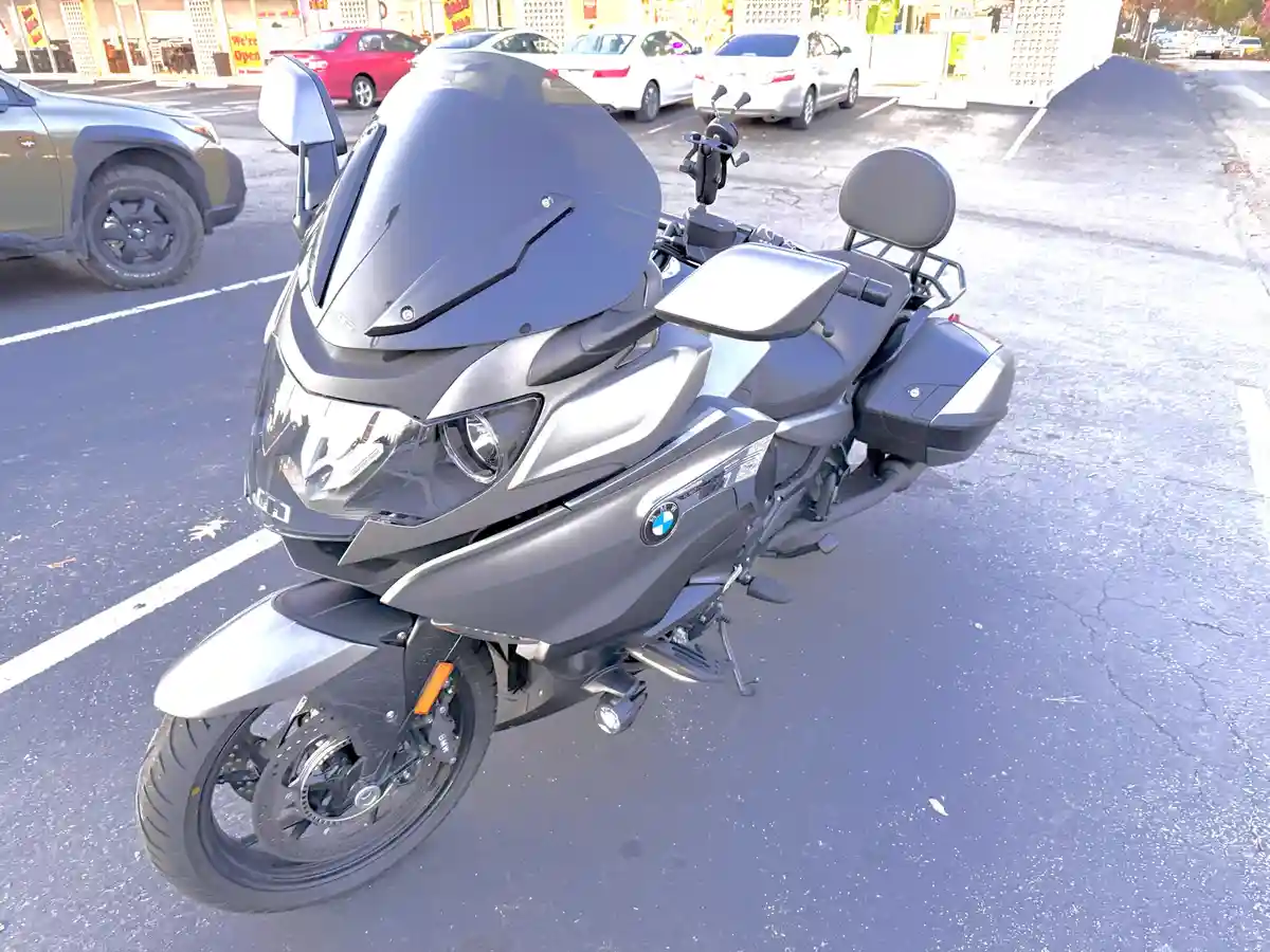 A silver BMW touring motorcycle is parked in a sunny parking lot, featuring a large windscreen, saddlebags, and a passenger…