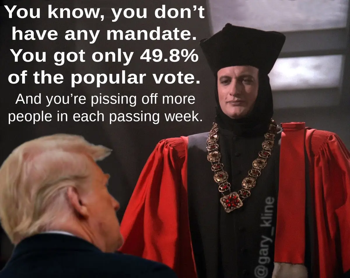 A man in a black hooded robe with a red cape and jeweled chain stands facing another man with blond hair seen from behind…