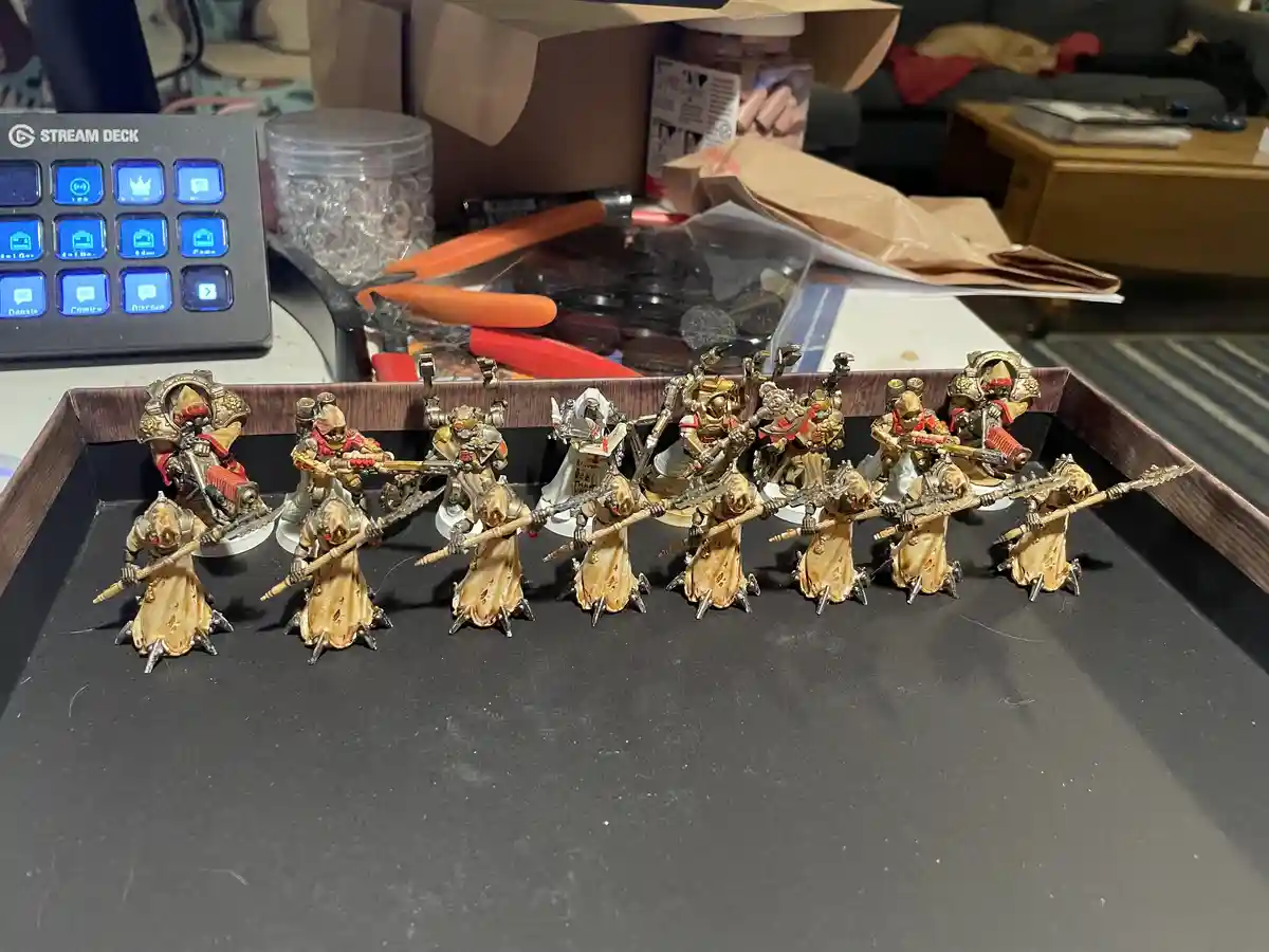 A row of painted miniature figurines, mostly in beige robes with some in ornate armor, are arranged on a black tray. Behind…