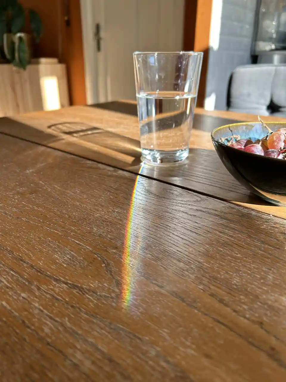 A glass of water sits on a wooden table, casting a rainbow prism effect on the surface due to sunlight refraction. Beside it…