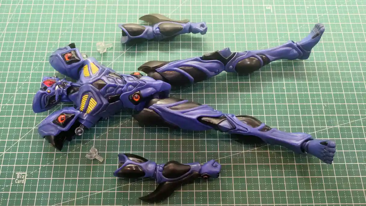 A blue and black plastic action figure is laid out on a green cutting mat, with its torso, head, and limbs separated and…