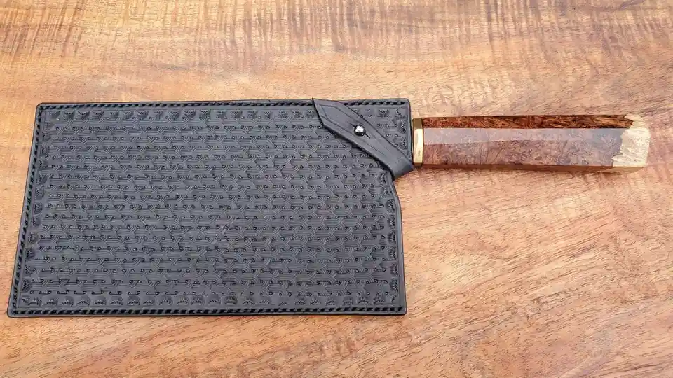 a knife with a decorative wooden handle and a black leather sheath lies on a wooden surface. The sheath is embossed with a…