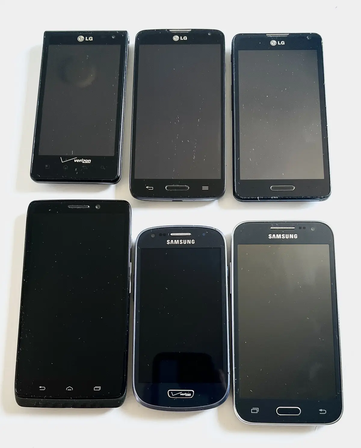 Six black smartphones, three LG and three Samsung models, are arranged in two rows on a white surface. All phones have dark…