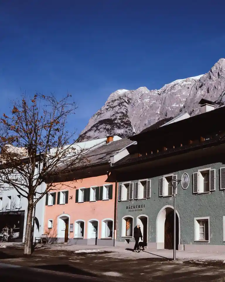 A picturesque street scene in a mountain town features colorful buildings with arched doorways and shuttered windows, set…