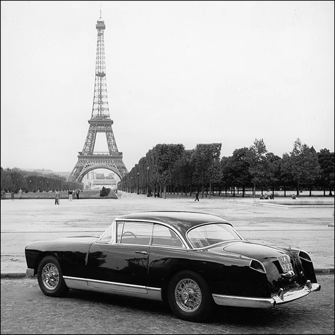 A black-and-white photograph shows a vintage, sleek black car parked in front of the Eiffel Tower in Paris. The car has a…