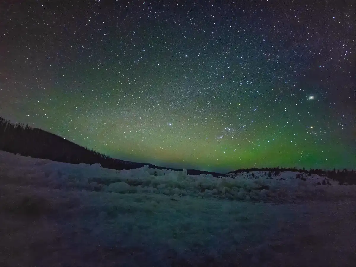 A stunning night sky photograph shows a vibrant green aurora borealis dancing across a star-filled expanse above a snowy…