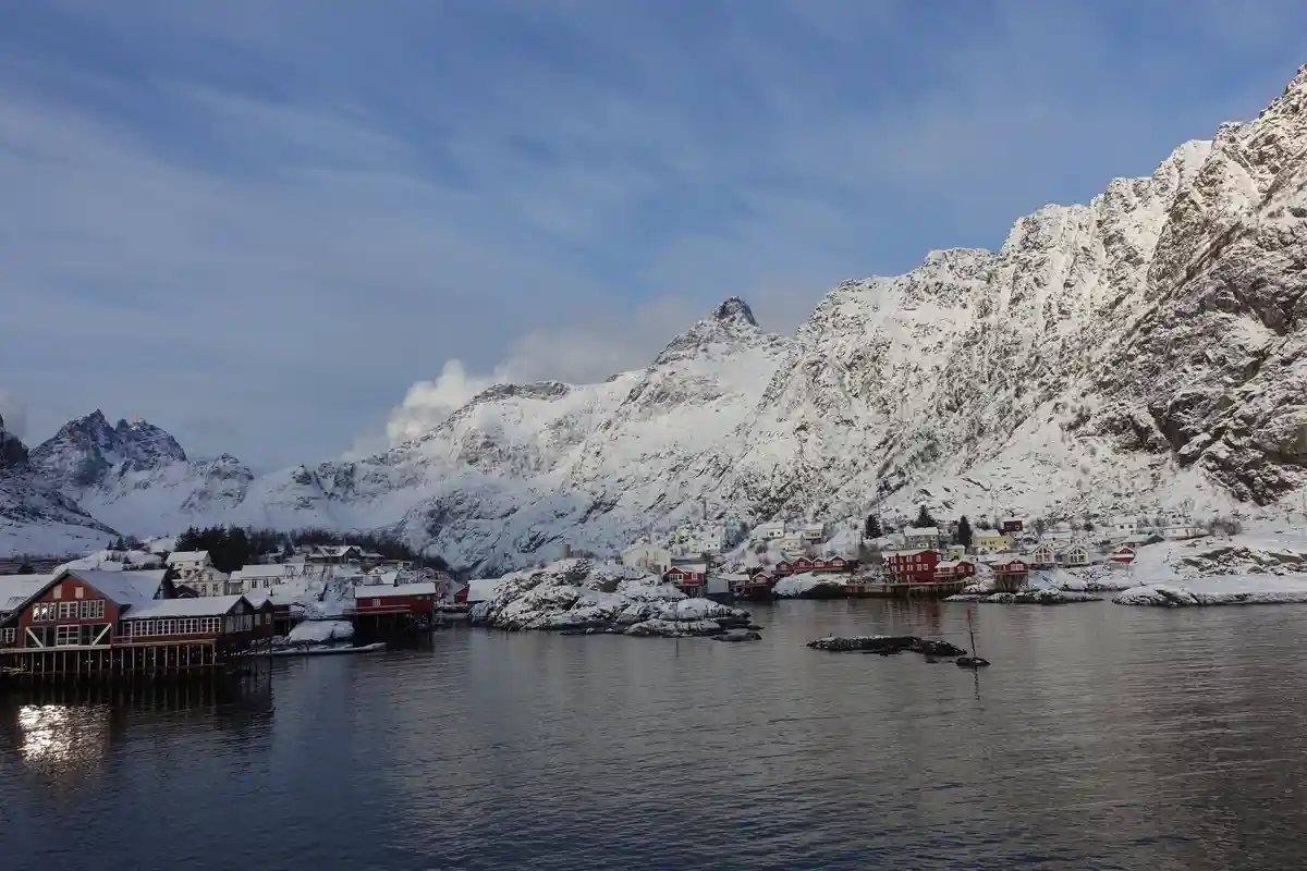 A scenic winter landscape shows a small village with red and white houses built along a calm body of water, surrounded by…