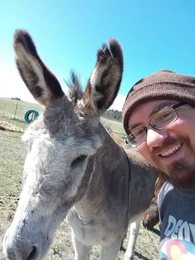 A selfie shows a smiling man with glasses and a brown beanie standing next to a donkey, both looking at the camera. The…