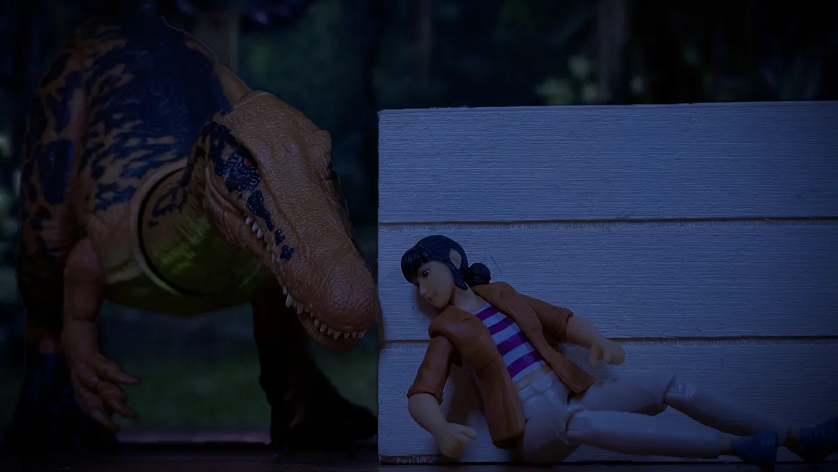 A toy dinosaur with a brown and black patterned body and red eyes looms over a toy human figure with black hair, a brown…