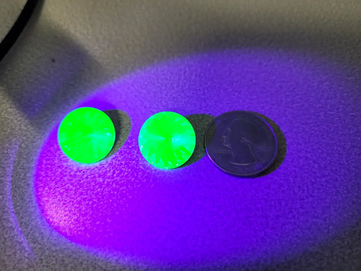 Under a purple UV light, two bright green, faceted gems glow intensely next to a United States quarter dollar coin on a…