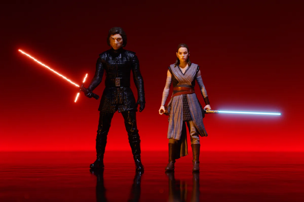 Two Star Wars action figures, Kylo Ren with a red crossguard lightsaber and Rey with a blue lightsaber, stand facing forward…